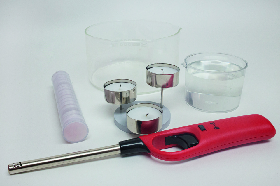 Im Vordergrund befinden sich ein roter Feuerzeug, drei Teelichter und eine transparente Wasserbecher. Daneben liegt ein r&ouml;hrenf&ouml;rmiger Beh&auml;lter und ein weiterer Messbecher mit klarem Wasser, die f&uuml;r Experimente im Bildungsbereich genutzt werden k&ouml;nnen.