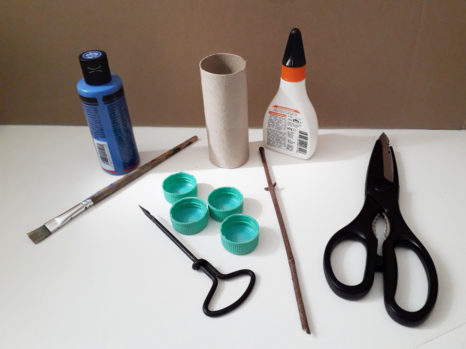 Auf dem Tisch liegen verschiedene Bastelmaterialien, darunter eine Flasche blauer Farbe, Kleber, eine Schere, ein Pinsel, ein Kartonrohr, mehrere Kunststoffdeckel, ein Stock und ein Werkzeug. Diese Utensilien eignen sich gut f&uuml;r kreative Experimente und Bastelprojekte im Bildungsbereich.