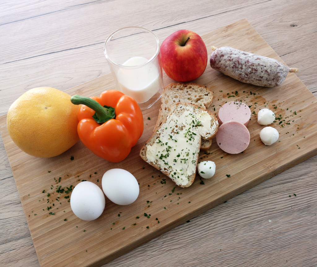 Auf einem Holzbrett sind verschiedene Lebensmittel sortiert: ein gelber Melon, eine orange Paprika, ein Apfel, mehrere Eier, Brot mit Butter, Wurstaufschnitt und Mozzarella-Bällchen. Neben den Lebensmitteln liegen frische Kräuter verstreut, was das Arrangement appetitlich aussehen lässt.
