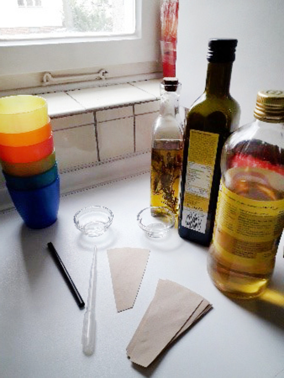 Farbenfrohe Becher stehen neben Flaschen mit verschiedenen Fl&uuml;ssigkeiten, darunter Oliven&ouml;l. Auf dem Tisch liegen zwei Papierst&uuml;cke, eine Pipette und zwei kleine Schalen, die auf ein Experiment hinweisen.