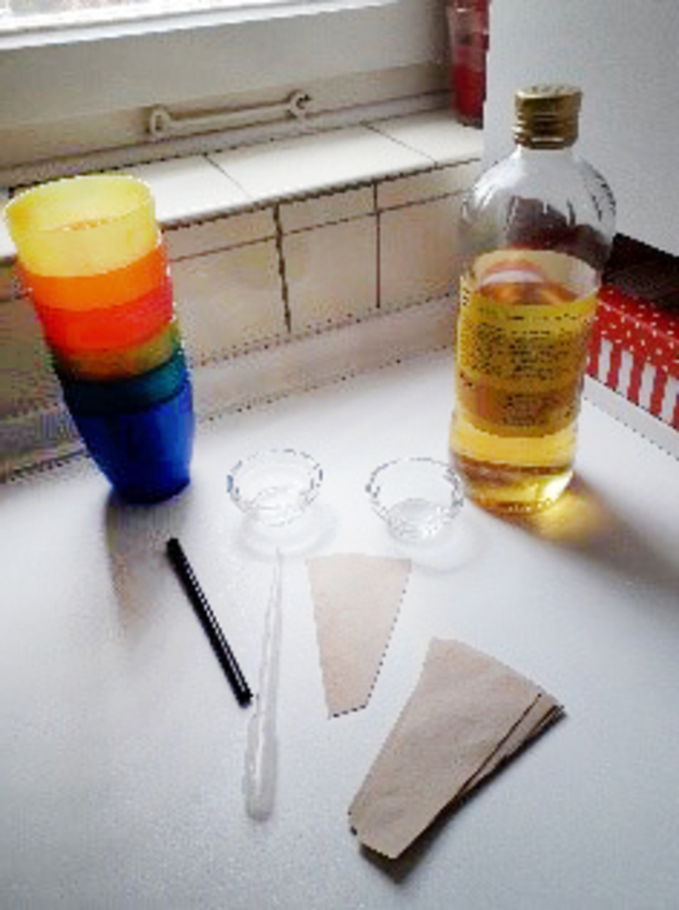 Bunte Becher aus Kunststoff stehen gestapelt neben einer Flasche mit Fl&uuml;ssigkeit und zwei kleinen Schalen. Au&szlig;erdem sind ein Strohhalm und zwei Papiert&uuml;ten auf dem Tisch verteilt, die f&uuml;r Experimente im Bildungsbereich verwendet werden k&ouml;nnen.