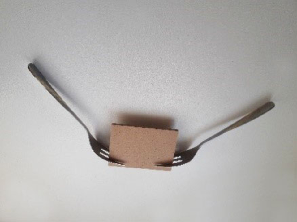 Zwei Gabeln sind an einem kleinen, quadratischen St&uuml;ck Karton befestigt, das in der Mitte des Bildes liegt. Die Gabeln ragen nach au&szlig;en und scheinen eine Art Halterung oder Unterst&uuml;tzung f&uuml;r das Kartonst&uuml;ck zu bilden.