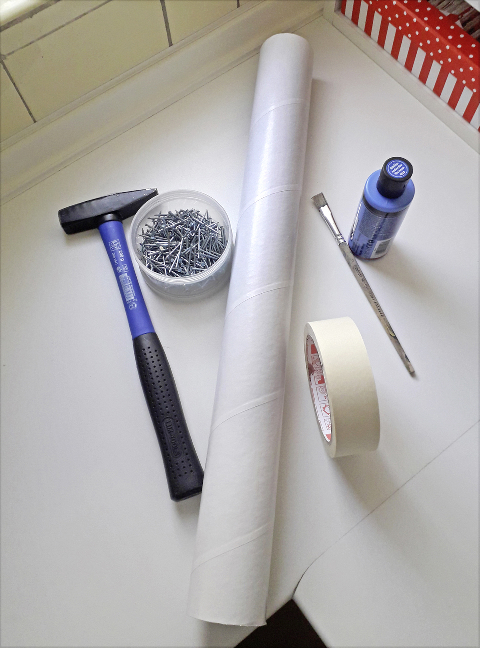 Auf der Arbeitsfl&auml;che sind verschiedene Materialien f&uuml;r experimentelle Projekte angeordnet. Zu sehen sind ein Hammer, eine Schachtel mit N&auml;geln, ein Rolle Papier, Buntfarbe samt Pinsel sowie Klebeband, alles f&uuml;r kreative Aktivit&auml;ten im Bildungsbereich.