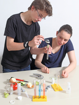Zwei Schüler führen ein Experiment durch, bei dem sie eine Probe mit einer Pipette in ein Reagenzglas übertragen. Auf dem Tisch sind verschiedene Obst- und Gemüsesorten sowie chemische Utensilien wie Reagenzgläser und Messbecher angeordnet.