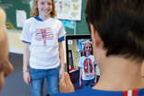 Eine Schülerin trägt ein T-Shirt mit einer anatomischen Darstellung, während ein Mitschüler mit einem Tablet ein digitales Modell ihrer Körperstruktur zeigt. Im Hintergrund ist eine Klassenraumkulisse mit einem überfüllten Mitteilungsbrett und interaktiven Elementen zu sehen.