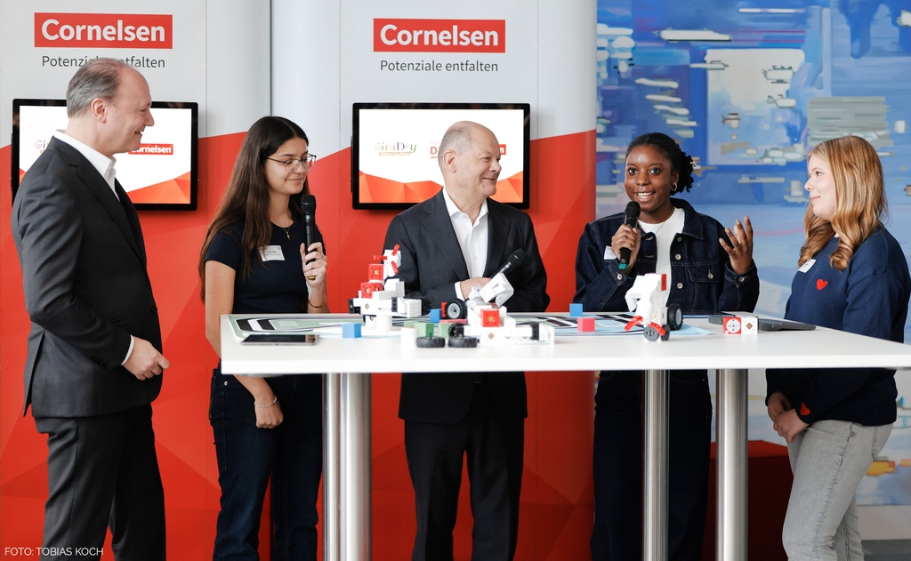 Fünf Personen stehen an einem Tisch und unterhalten sich mit Mikrofonen. Auf dem Tisch sind Lernroboter aus dem Bildungsbereich zu sehen, während im Hintergrund ein farbiger Banner von Cornelsen abgebildet ist.