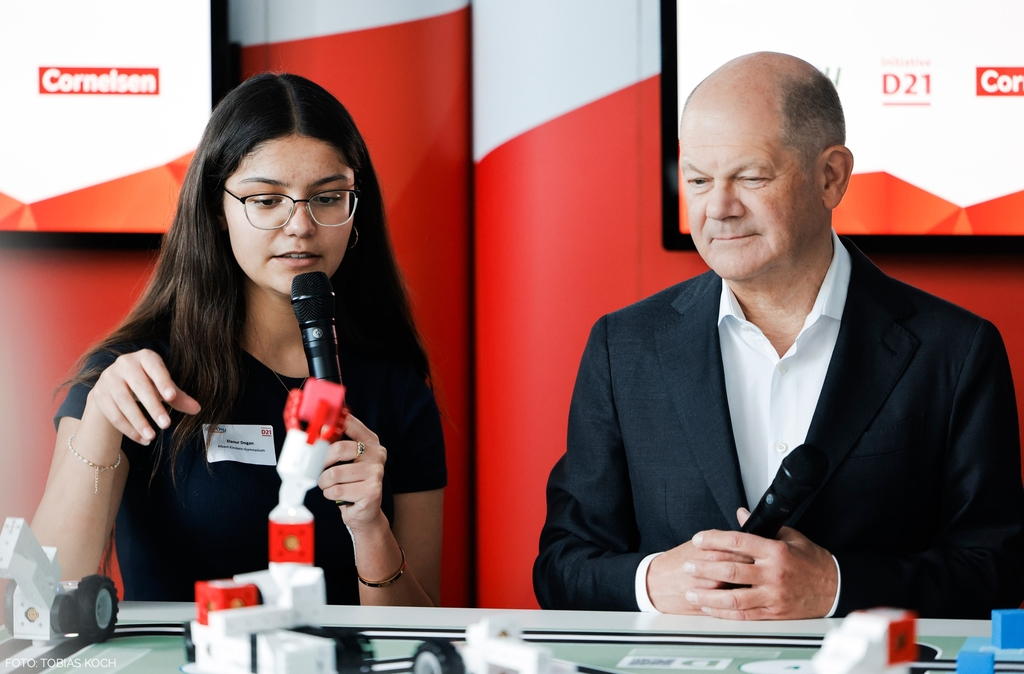 Eine junge Frau pr&auml;sentiert den Lernroboter eXperiBot mit einem Mikrofon in der Hand, w&auml;hrend Bundeskanzler Scholz aufmerksam zuschaut. Im Hintergrund sind Logos und eine kreative Umgebung sichtbar, die auf eine Bildungs- und Innovationsveranstaltung hinweisen.