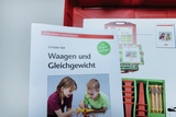 Ein Schüler-Set für Experimente zu Waagen und Gleichgewicht ist abgebildet. Es umfasst eine Anleitung mit Kopiervorlagen und zeigt anschauliche Materialien sowie Aufnahmen von Kindern, die mit den Experimenten arbeiten.