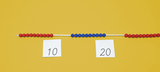 Auf einer gelben Unterlage liegen zwei Karten mit den Zahlen 10 und 20. Zwischen den Karten verläuft eine Schnur, an der rote und blaue Perlen aufgereiht sind, wobei die rote Perlenreihe auf der linken Seite und die blaue auf der rechten Seite platziert ist.