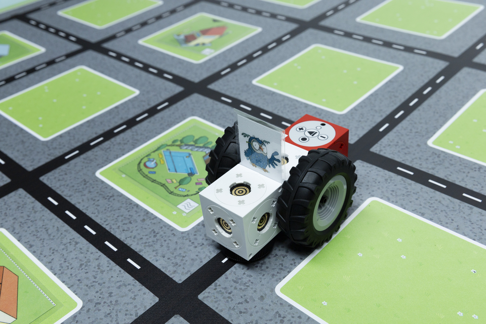 Ein fahrzeug&auml;hnliches Modell mit gro&szlig;en Reifen steht auf einem matten Untergrund, der Stra&szlig;en und gr&uuml;ne Bereiche darstellt. Das Modell zeigt bunte W&uuml;rfel mit Symbolen und einem Bild eines Vogels auf der R&uuml;ckseite.