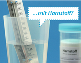 Im Bild ist eintransparentes Gefäß mit Wasser und einem Thermometer zu sehen, das in das Wasser eingetaucht ist. Neben dem Gefäß steht ein Behälter mit der Aufschrift "Harnstoff", was auf mögliche chemische Experimente hinweist.