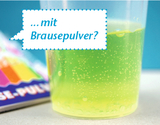 Im Vordergrund ist ein transparentes Becherglas mit einer sprudelnden, grünlichen Flüssigkeit zu sehen. Im Hintergrund liegt eine bunte Verpackung von Brausepulver, die auf eine mögliche Experimentieranleitung hindeutet.