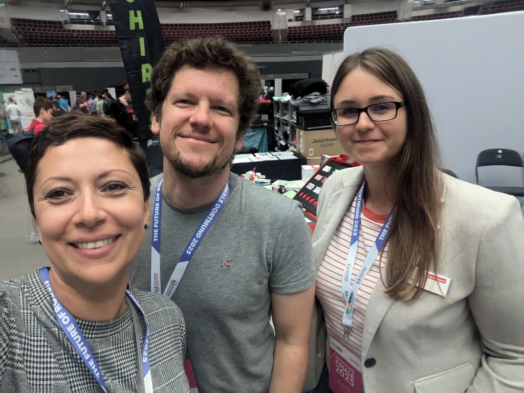 Drei Personen l&auml;cheln in die Kamera und scheinen an einem Stand bei einer Veranstaltung f&uuml;r Experimentierbedarf beteiligt zu sein. Im Hintergrund sind St&auml;nde und verschiedene Produkte zu erkennen, die f&uuml;r Bildungszwecke angeboten werden.