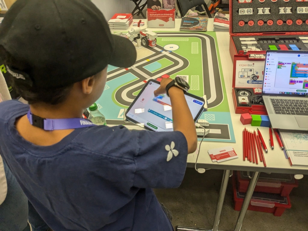 Ein Kind interagiert mit einem Tablet, um den eXperiBot Lernroboter zu steuern. Neben dem Tablet ist ein aufgeklappter Koffer des eXperiBot Smart Teams zu sehen sowie eine Spielmatte und darauf fahrende Lernroboter.
