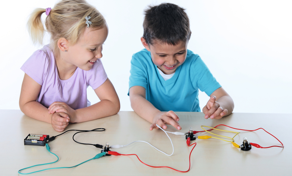 Zwei Kinder experimentieren am Tisch mit elektrischen Schaltkreisen. Sie verbinden Kabel zu Lampen und sind sichtbar begeistert von den Ergebnissen ihrer Experimente.