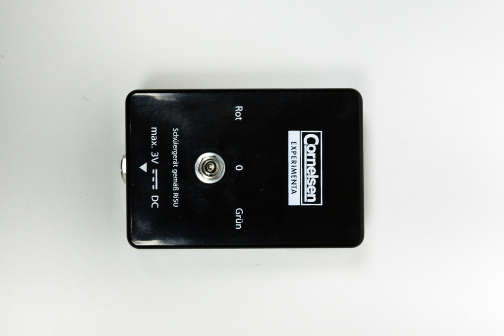 Ein schwarzes Experimentiergerät zeigt eine Anschlussbuchse sowie farbige Beschriftungen in Rot und Grün. Es ist für den Gebrauch in der Schule konzipiert und unterstützt eine maximale Spannung von 3 Volt Gleichstrom.