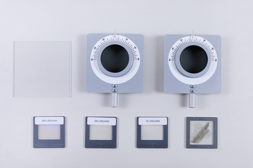 Zwei optische Geräte mit Skalen sind auf dem Bild zu sehen, daneben liegt ein Glasplättchen. Unterhalb der Geräte befinden sich drei verschiedene Folien mit einer Kennzeichnung für die Anzahl der Linien pro Millimeter.