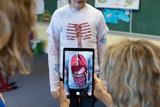 Ein Kind trägt ein T-Shirt mit einer Darstellung des menschlichen Skeletts und innerer Organe. Eine Person hält ein Tablet, das eine digitale Überlagerung der Organe zeigt, um den Lehrinhalt interaktiv zu vermitteln.