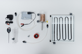 Auf der weißen Unterlage sind verschiedene Komponenten eines Experimentierkits angeordnet, darunter ein Heizelement, Schläuche, ein Thermometer und ein Wasserbehälter. Diese Teile sind für technische Experimente im Bildungsbereich konzipiert und dienen der Veranschaulichung von thermodynamischen Prozessen.