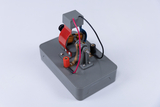 Ein experimentelles Gerät zur Demonstration elektrischer Prinzipien ist abgebildet, das mit verschiedenen Kabeln und Anschlüssen ausgestattet ist. Es zeigt eine Anordnung aus Spulen und mechanischen Komponenten, die zur Erzeugung von Bewegung oder Strom verwendet werden kann.
