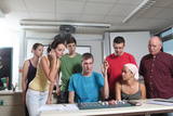 Eine Gruppe von sieben Personen, darunter mehrere Jugendliche und ein älterer Mann, arbeiten gemeinsam an einem Experiment in einem Klassenzimmer. Der Schwerpunkt liegt auf einem Tisch, auf dem ein Experimentiergerät zu sehen ist, während die Teilnehmer aufmerksam eine Demonstration verfolgen.