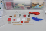 Verschiedene Alltagsgegenstände wie eine Feder, Steine, Papier und eine Pipette sind ordentlich auf einer Fläche ausgelegt. Im Hintergrund befindet sich eine Kunststoffbox mit weiteren Utensilien wie Markern und Knete.