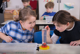 Zwei Mädchen führen konzentriert ein wissenschaftliches Experiment mit einem Messzylinder und Pipetten durch. Im Hintergrund sind weitere Schüler in einem Klassenzimmer zu sehen.