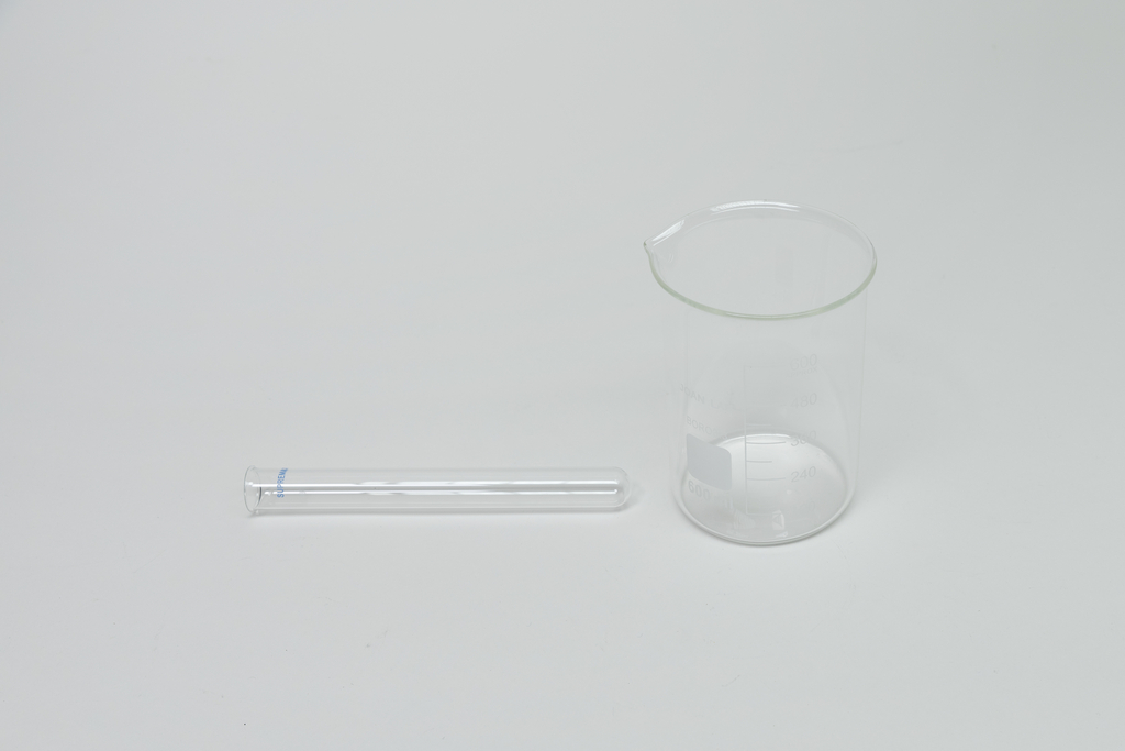 Ein durchsichtiges Becherglas aus Glas steht auf einer weißen Oberfläche. Daneben liegt ein passender Glasdeckel.