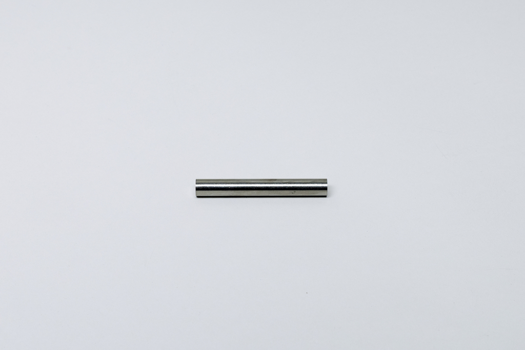Ein kleiner, zylindrischer Metallstab liegt horizontal auf einem hellgrauen Untergrund. Er weist eine glatte, reflektierende Oberfläche auf.