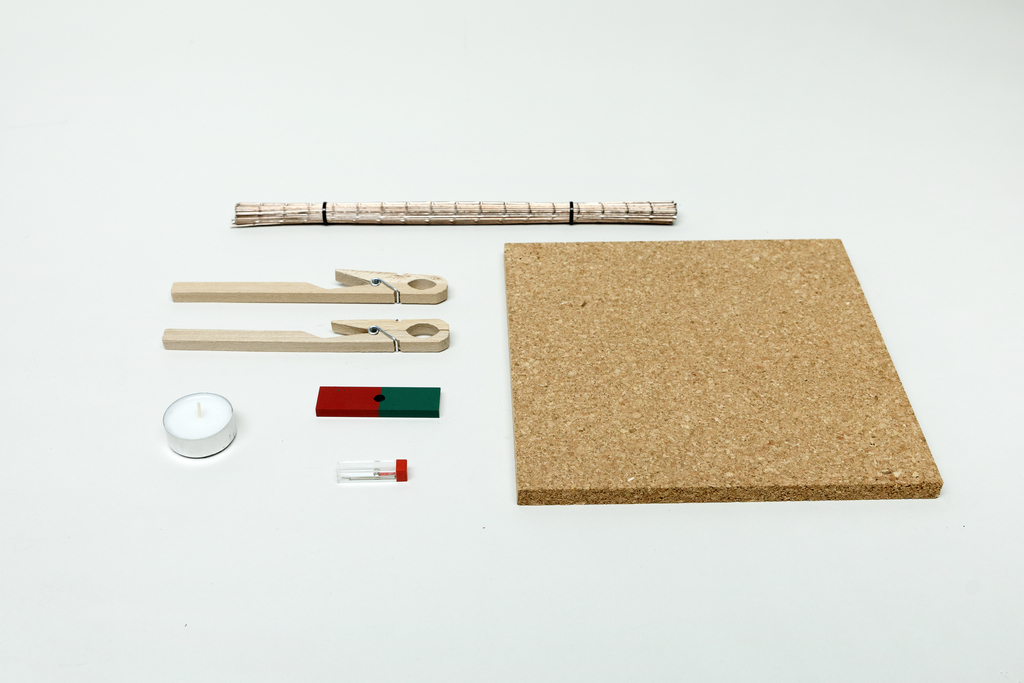 Ein Set aus Experimentierbedarf umfasst eine Korkplatte, Holzklammern und ein Bündel Strohhalme. Ergänzt wird das Ensemble durch ein Teelicht, ein kleines Reagenzglas und einen zweifarbigen Holzblock.