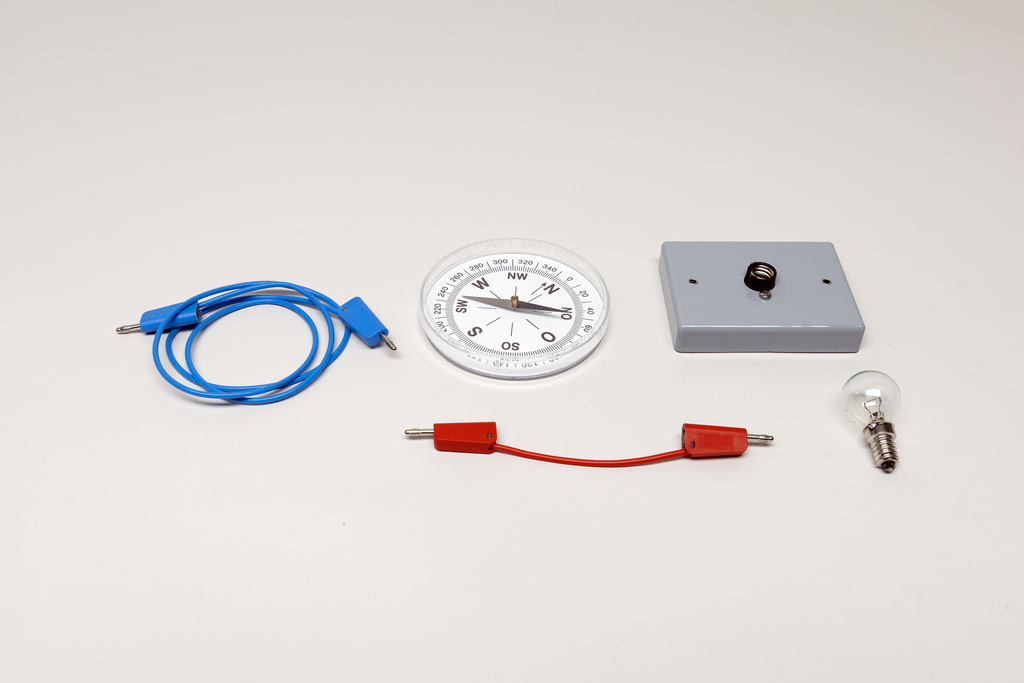 Ein Kompass, eine Glühbirne und ein grauer Schaltblock liegen auf weißem Grund. Zwei Anschlusskabel in Blau und Rot ergänzen das Set für physikalische Experimente.