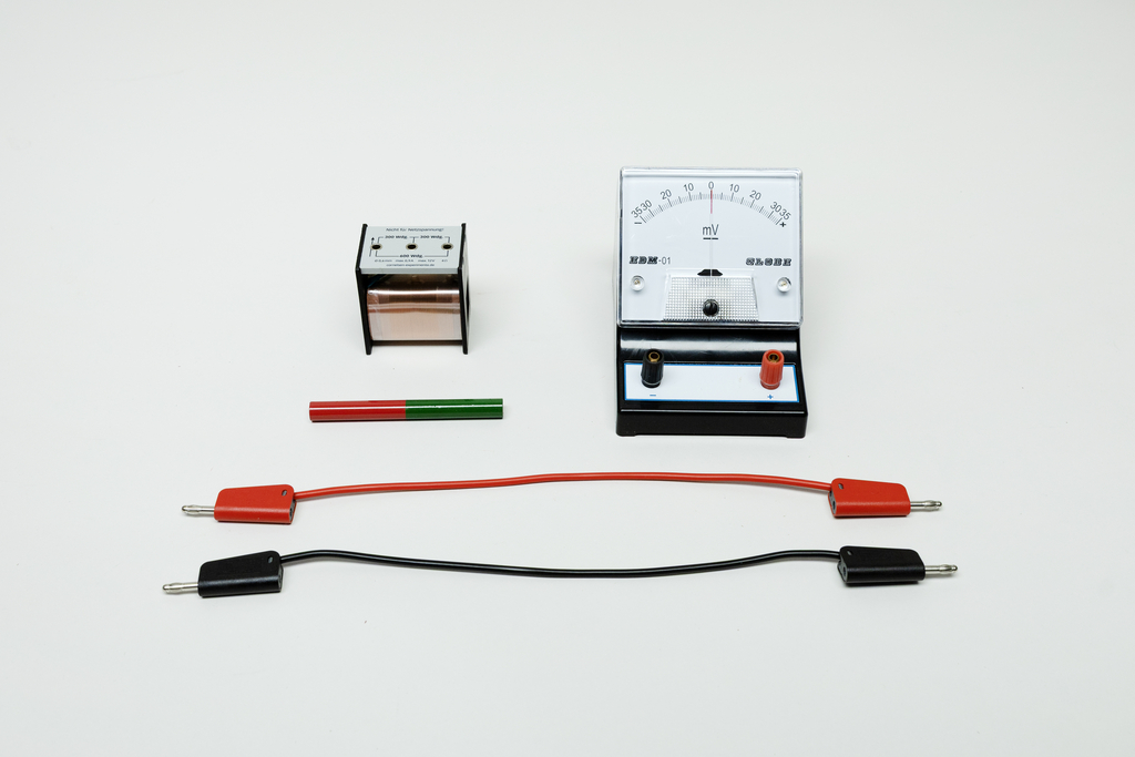 Ein Millivoltmeter, ein Spulenkörper mit Eisenkern und zwei Anschlusskabel in Rot und Schwarz liegen auf weißem Grund. Diese Komponenten dienen physikalischen Experimenten zur Elektrizität.