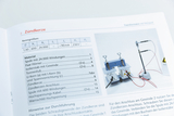 Ein offenes Heft zeigt eine Anleitung zum Thema Zündkerze inklusive einer Tabelle mit Kenngrößen. Rechts daneben ist ein Foto eines physikalischen Versuchsaufbaus mit Spulen und Kabeln abgebildet.