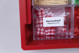 Ein roter Organizer enthält verschiedene Kunststoffbehälter mit Materialien. Eines der Fächer ist mit rot-weiß kariertem Baumwollstoff gefüllt und entsprechend beschriftet.