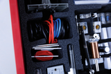 Ein roter Koffer enthält in Schaumstoffaussparungen verschiedene technische Bauteile. Sichtbar sind unter anderem farbige Kabel, Spulen und elektrische Komponenten für Experimente.