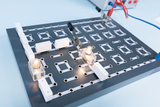 Ein elektrischer Schaltkreis ist auf einer grauen Versuchsplatte mit kleinen Glühlampen und weißen Verbindungsstücken aufgebaut. Über Krokodilklemmen ist das System mit einer externen Stromquelle verbunden.