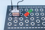 Ein elektrischer Transformator ist auf einer grauen Lochplatte montiert und mit Kabeln verbunden. Ein rot-grüner Magnet schwingt in dessen Nähe, um physikalische Prinzipien zu demonstrieren.