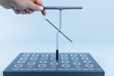 Eine Hand hält ein metallisches Gestell über eine quadratische Basisplatte mit einem Gitter aus elektrischen Kontakten. Es handelt sich um ein physikalisches Experimentierset zur Veranschaulichung elektrischer Stromkreise.