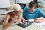 Zwei junge Menschen arbeiten gemeinsam an einem physikalischen Experiment mit einem elektronischen Baukasten. Sie konzentrieren sich auf die Schaltung auf einer schwarzen Basisplatte und machen Notizen in einem Heft.
