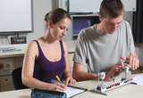 Zwei Jugendliche führen an einem Tisch ein physikalisches Experiment mit einer Luftkissenbahn durch. Während der Junge den Versuchsaufbau bedient, hält die Jugendliche die Ergebnisse schriftlich fest.