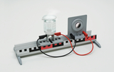 Ein physikalisches Experimentierset zeigt ein Becherglas und einen Spiegel auf einer Metallschiene. Rote und schwarze Kabel verbinden die Komponenten für eine optische oder elektrische Messung.