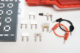 Ein Set aus elektrischen Bauteilen, wie Steckbrücken, Schaltern und Kabeln, ist auf einer weißen Fläche angeordnet. Daneben befindet sich ein graues Schaltbrett sowie ein roter Transportkoffer.