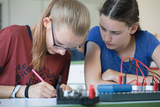 Zwei Schülerinnen arbeiten konzentriert an einem physikalischen Experiment mit einem Breadboard und Kabeln. Eine der Jugendlichen macht sich dabei Notizen auf einem Blatt Papier.