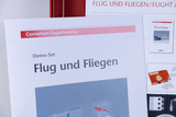 Eine Broschüre für ein Cornelsen Experimenta Demo-Set zum Thema „Flug und Fliegen“ steht im Vordergrund. Im Hintergrund ist ein weiteres Informationsblatt mit ähnlichem Inhalt sichtbar.
