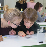 Zwei Schüler betrachten konzentriert eine Probe in einem kleinen Kunststoffbehälter auf einem Labortisch. Im Hintergrund sind weitere Mitschüler in einem hellen Klassenzimmer zu sehen.