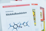 Eine Handreichung für Molekülbaukästen von Cornelsen Experimenta liegt im Vordergrund. Im Hintergrund sind weitere Unterlagen und ein roter Koffer mit Chemie-Equipment zu sehen.