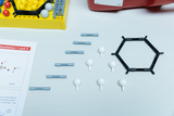 Ein Chemie-Molekülbaukasten mit grauen Verbindungsstücken und weißen Atomen liegt auf einer weißen Fläche. Daneben befinden sich eine Anleitung sowie eine gelbe Sortierbox mit weiteren Bauteilen.