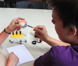 Ein Schüler führt ein chemisches Experiment durch und vergleicht eine Teststreifenfarbe mit einer Farbskala. Vor ihm auf dem Tisch befinden sich zudem Reagenzgläser in einem Ständer, ein Becherglas und Notizmaterial.