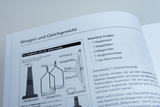 Ein offenes Lehrbuch zeigt eine Anleitung zum Zusammenbau einer Balkenwaage inklusive einer Materialliste. Schematische Zeichnungen veranschaulichen die einzelnen Komponenten wie Waagebalken und Waagschalen.